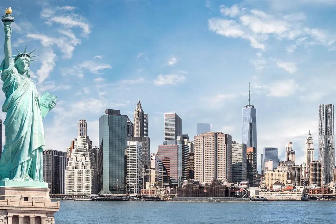 The Statue of Liberty in front of the Manhattan skyline on a clear day.