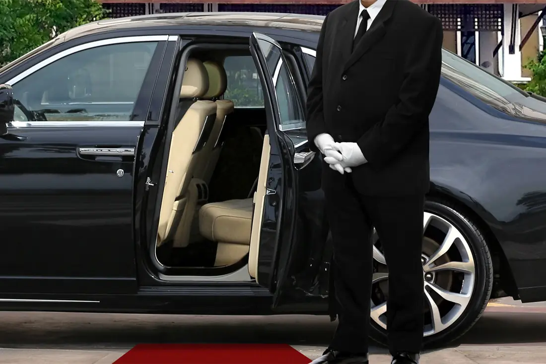 A driver in a dark suit holding open the door of a black car.