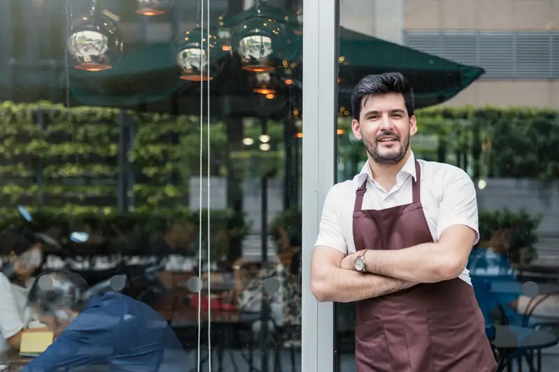 A café owner in his apron, arms folded, outside his front window.
