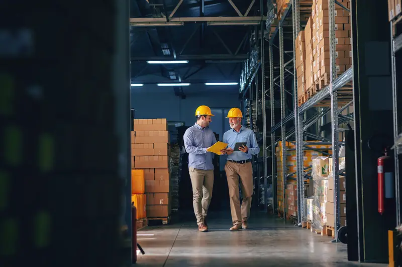 Two warehouse managers in hard hats walking an aisle of stacked pallets.